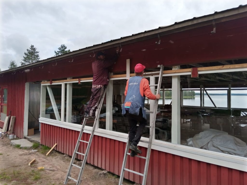 Kuvassa kaksi miestä kiipeää tikapuille ja rakentaa kattoa. 